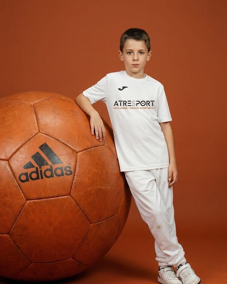 Un niño con la camiseta blanca de ATRESPORT, de pie junto a un balón de cuero.